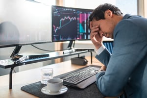 An investor touches his head in frustration while sitting at a desk in front of a computer and a cup of coffee.